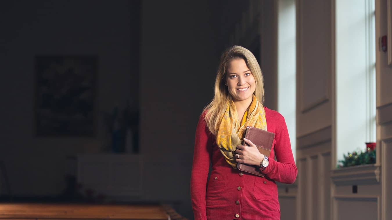 An alumna of the MDiv in Theology & Ministry - Church and Ministry degree program at Regent University standing in the chapel on campus holding a Bible and smiling