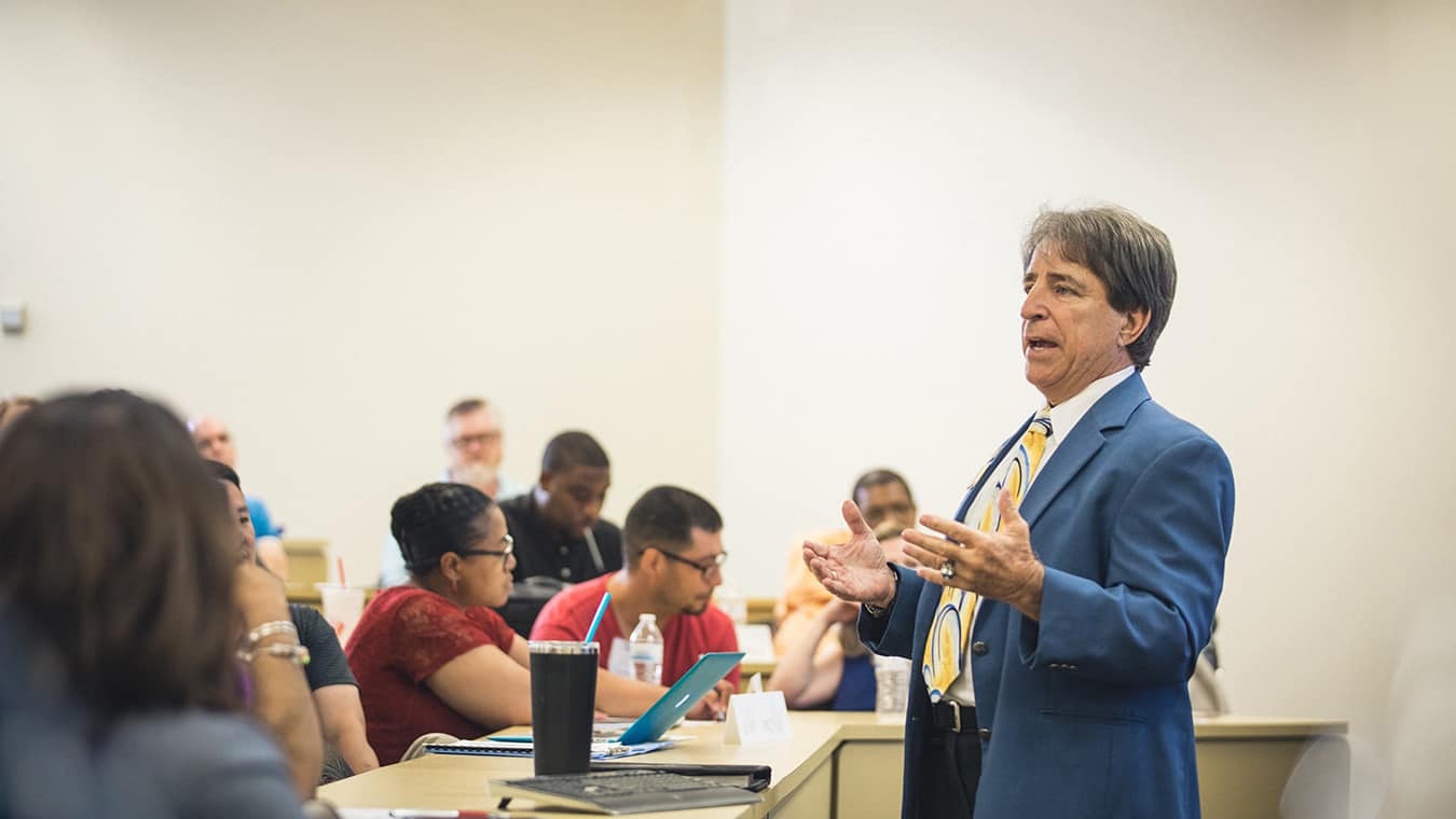 A professor teaching a class with a row of students in Regent's Ph.D. Higher Education online program.