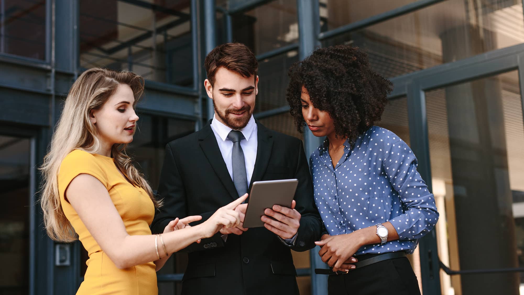 Two women and one man, dressed in business attire, standing outside a modern office building and looking at a tablet together.